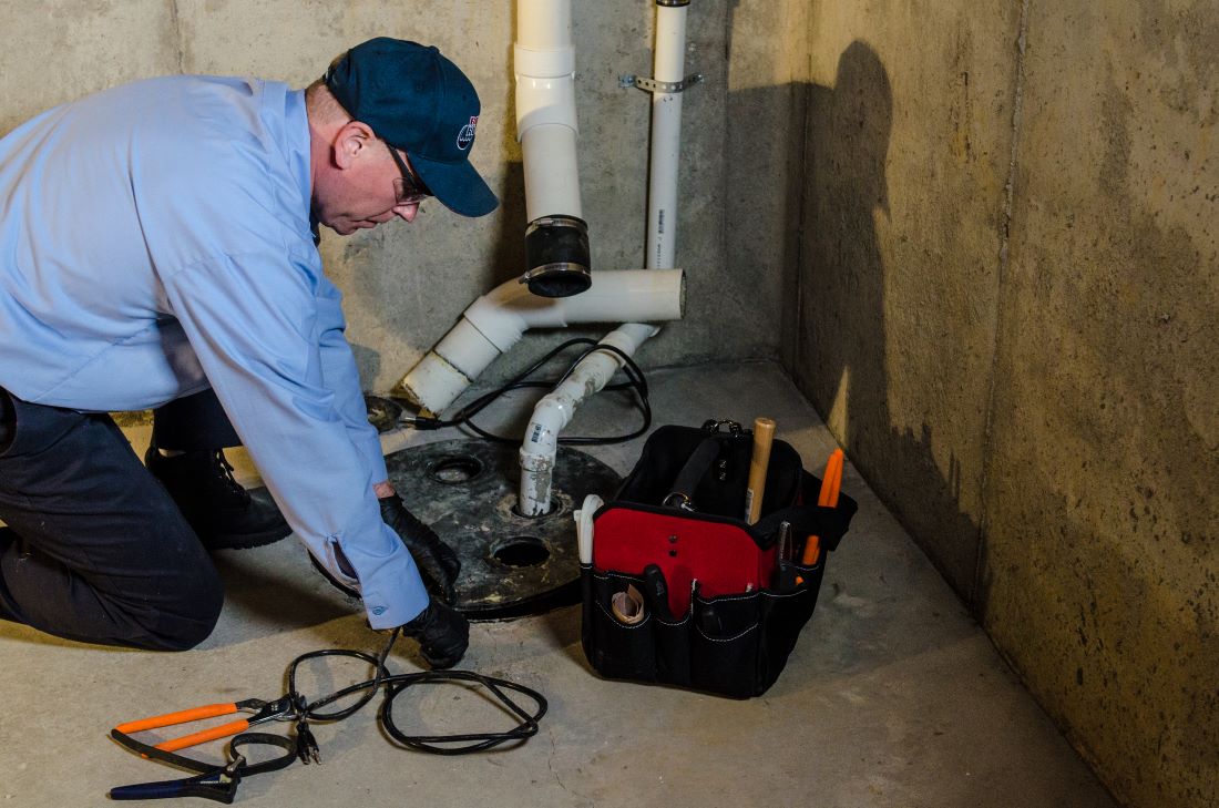 Plumber evaluating sump pump while kneeling on ground next to tool box and sump pit